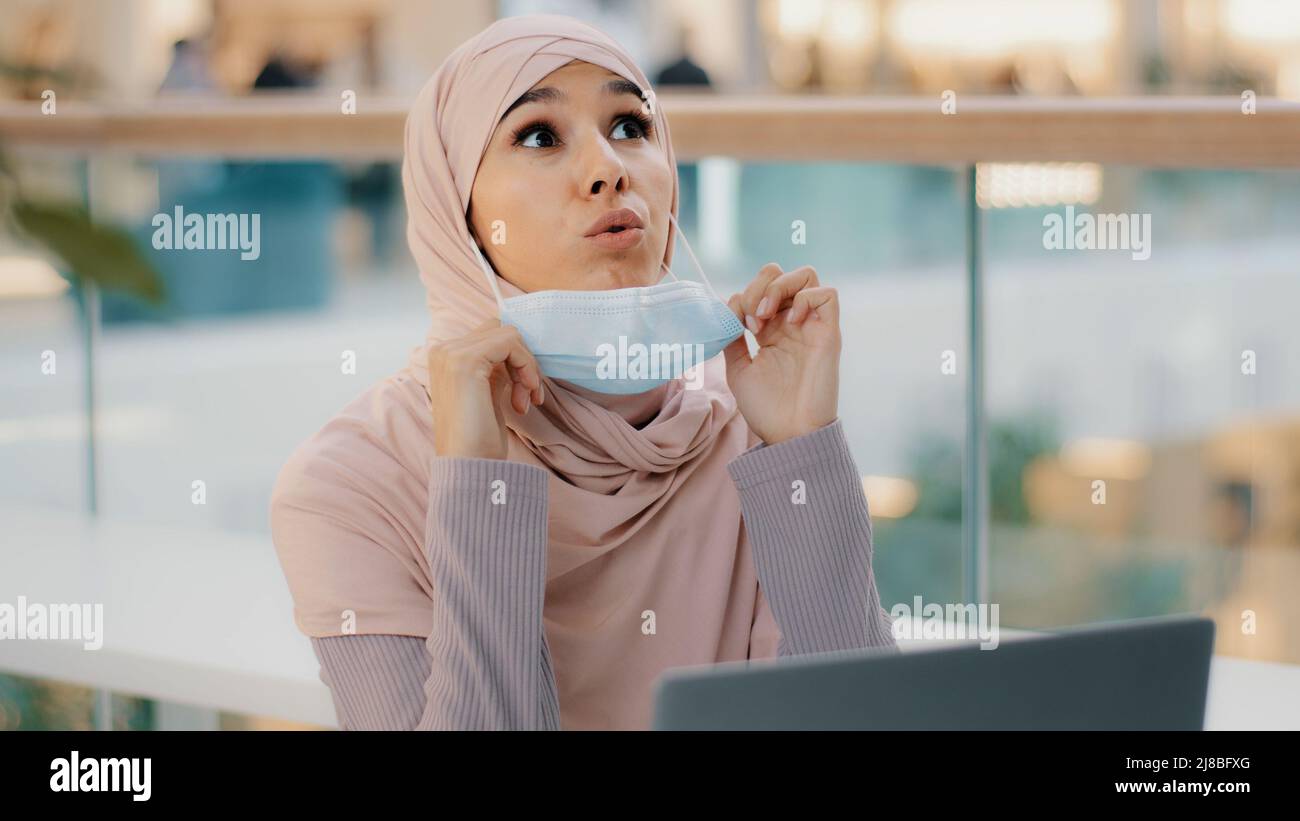 Giovane donna d'affari araba in hijab lavora a laptop guarda intorno musulmana ragazza abbassa maschera medica espira con rilievo divertente graffi naso mette Foto Stock