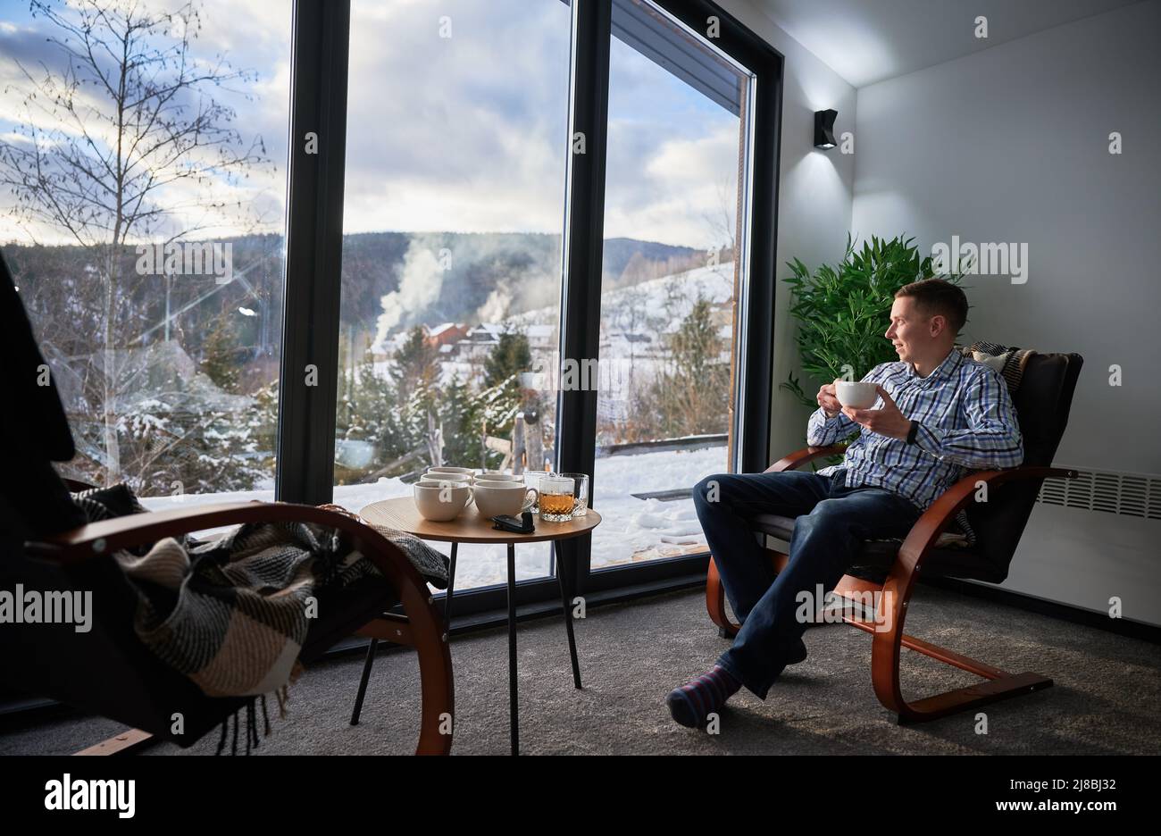 Giovane uomo che si gode week-end all'interno di una caserma contemporanea. Felice turista maschile seduta su sedia, tenendo una tazza di tè, godendo di vista del paesaggio di montagna attraverso finestre panoramiche in nuovo cottage. Foto Stock