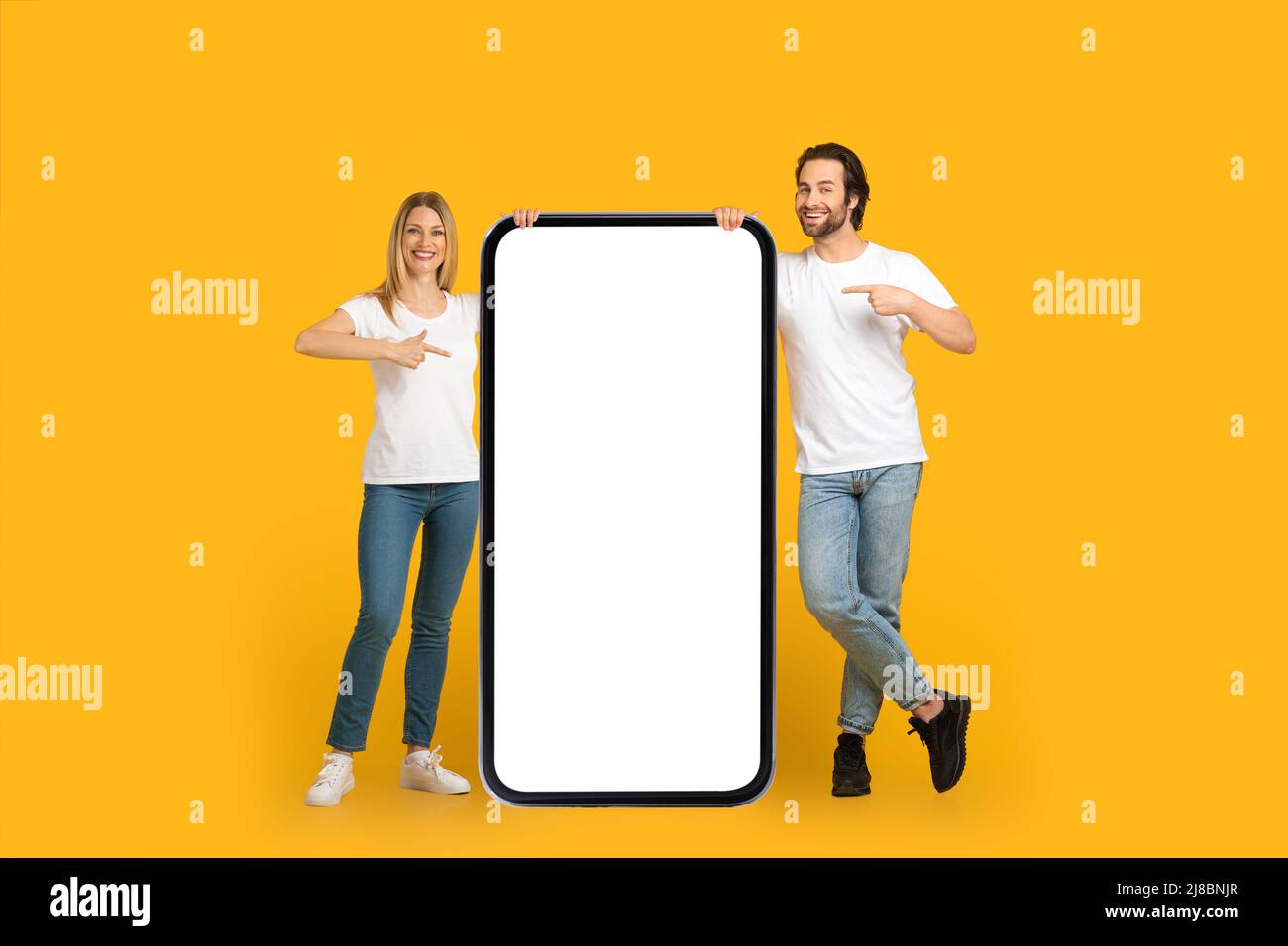 Soddisfatto coppia millennial europea in magliette bianche puntano le dita su un telefono enorme con schermo vuoto Foto Stock