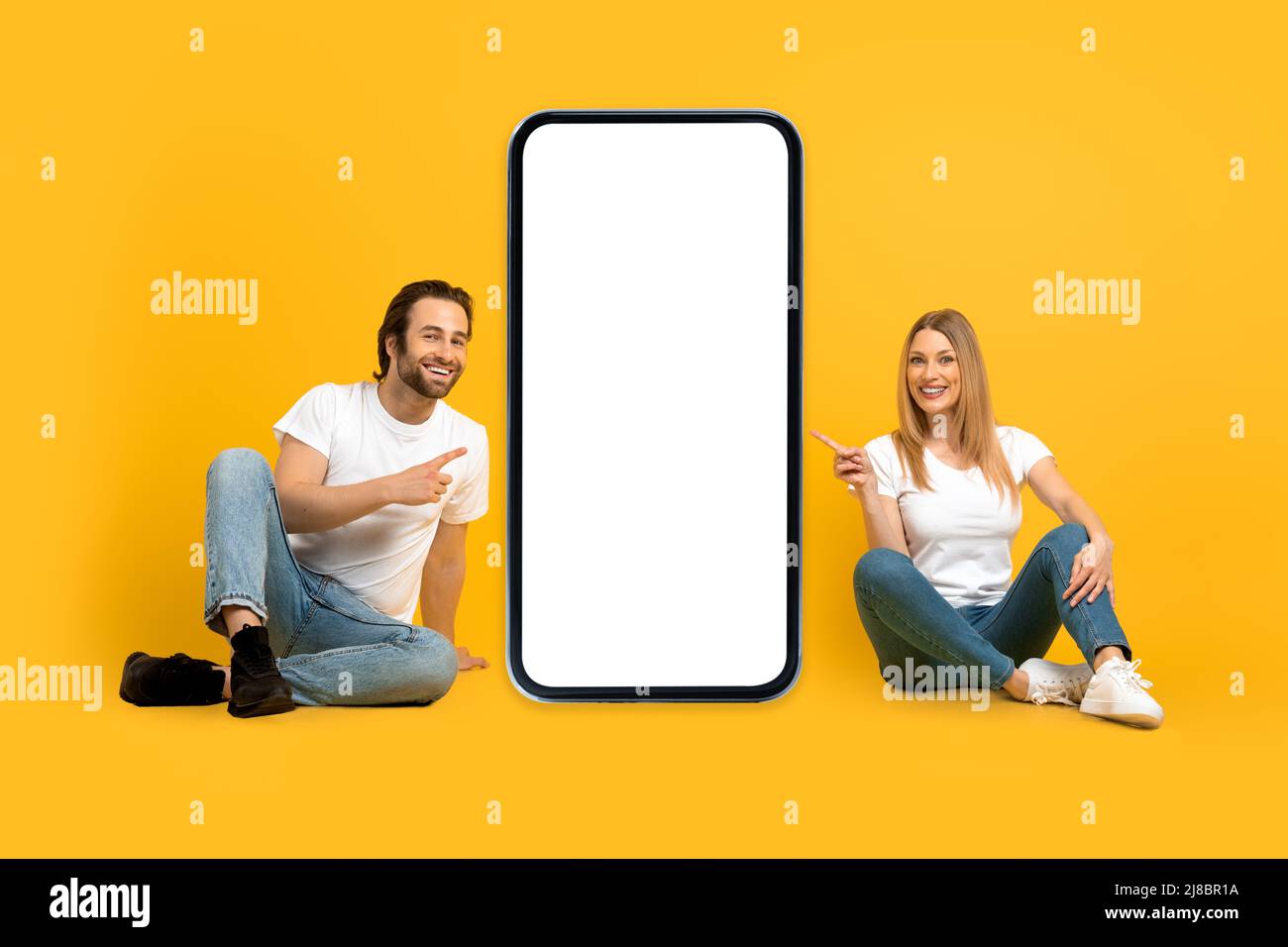 Felice giovane moglie europea e marito con stoppia sedersi a terra, puntare le dita verso il grande smartphone con schermo vuoto Foto Stock