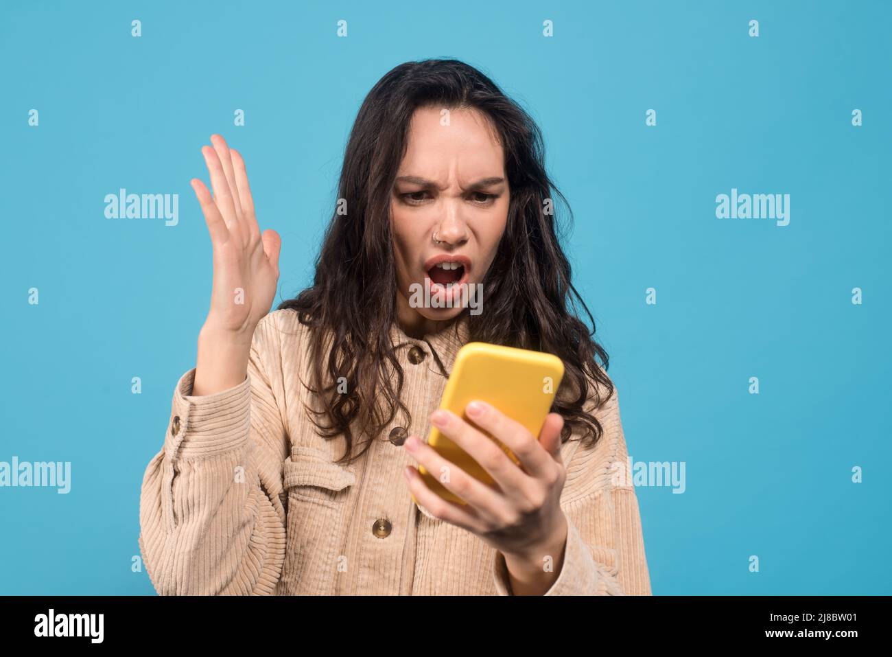 Triste scioccato giovane donna bruna europea con bocca aperta guarda smartphone isolato su sfondo blu Foto Stock