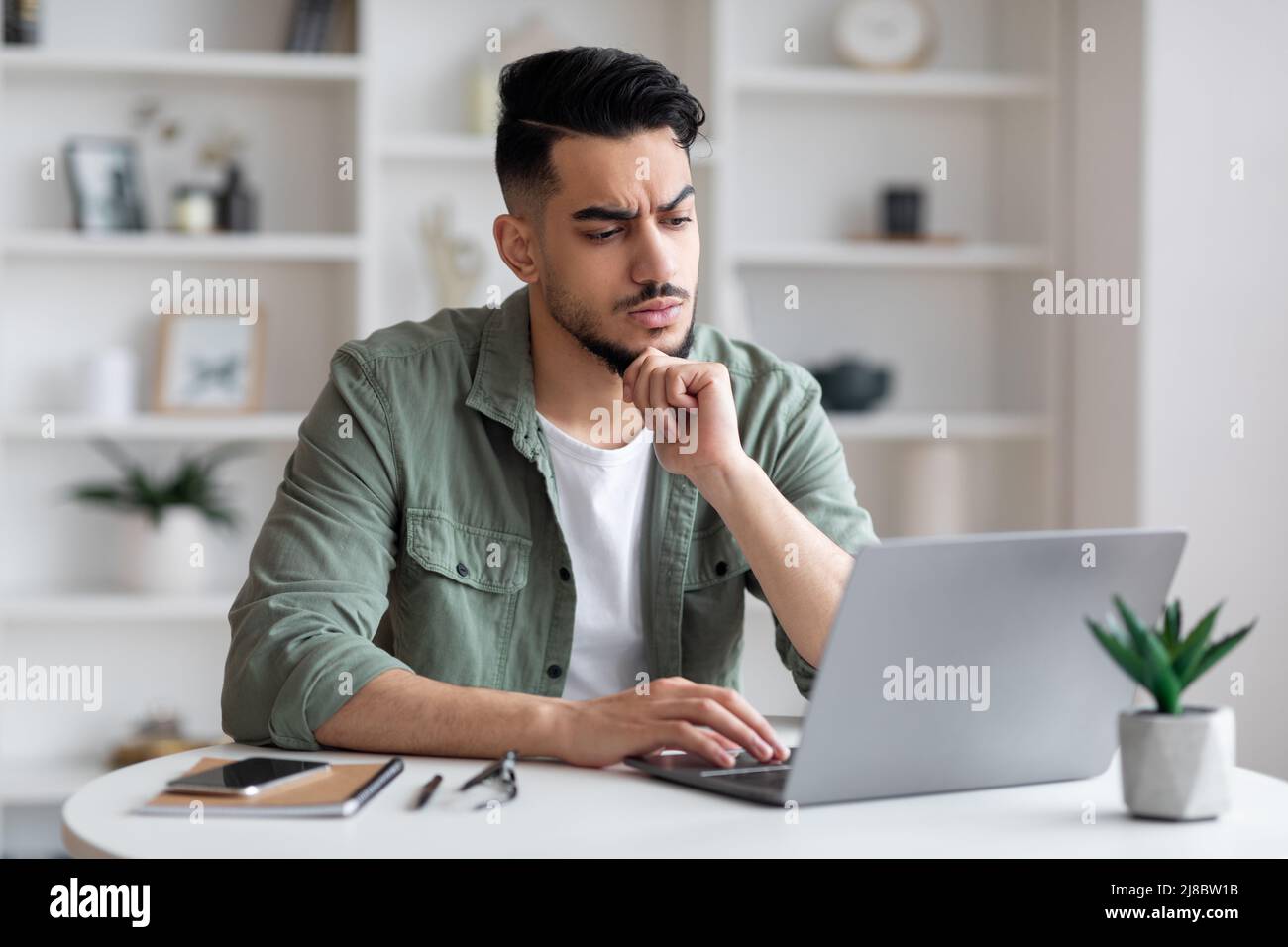 Il giovane e triste uomo arabo, concentrato e premuroso, con la barba che batte sul computer portatile e ha problemi con il lavoro Foto Stock