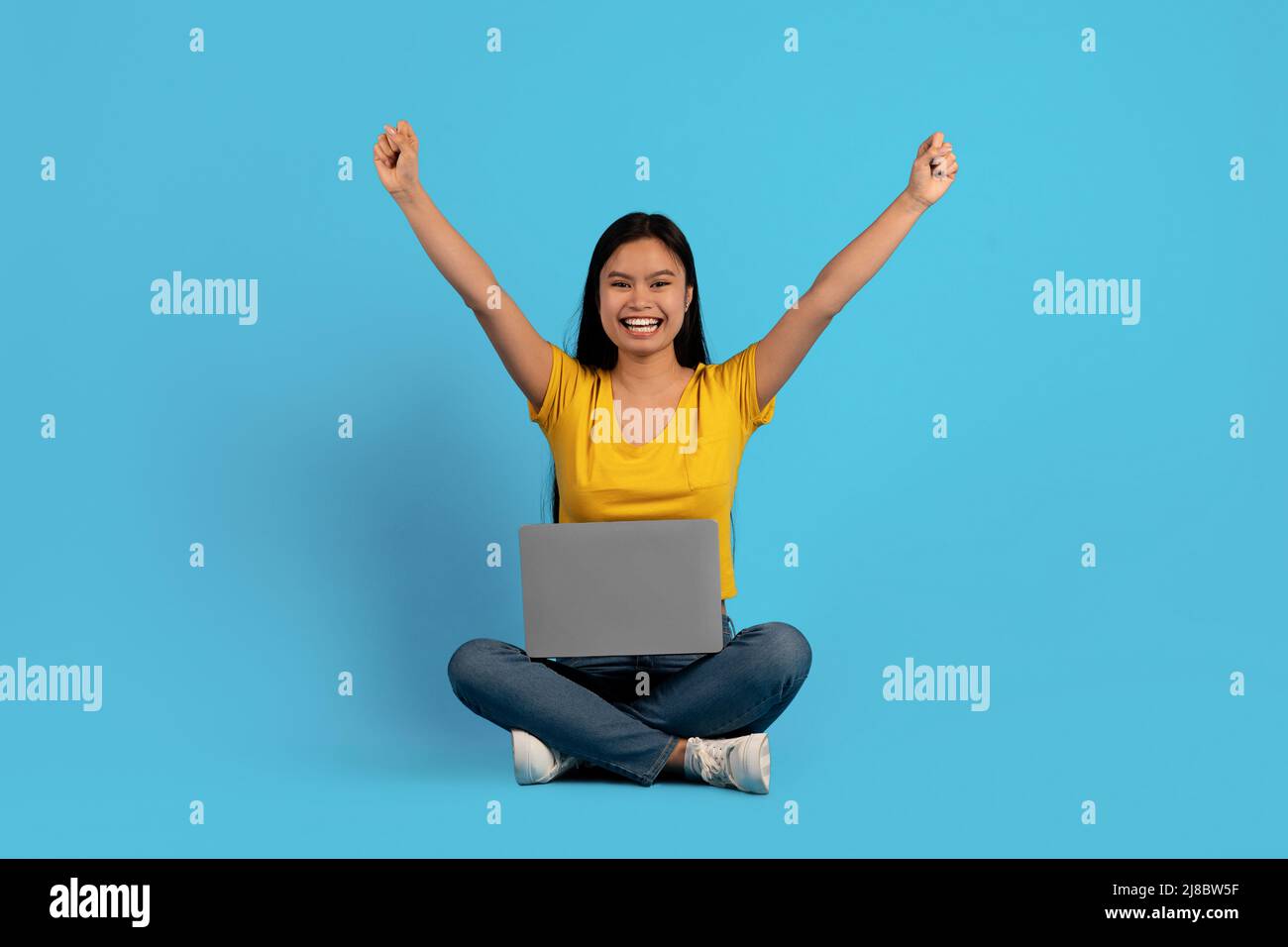 Felice giovane donna cinese in casual si siede sul pavimento con il computer portatile e solleva le mani in su gioisce nella vittoria Foto Stock