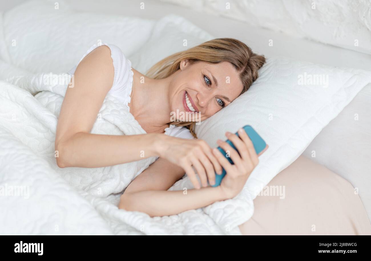 Felice bella giovane bionda europea donna legge messaggio al telefono, sdraiato sul letto in camera bianca Foto Stock