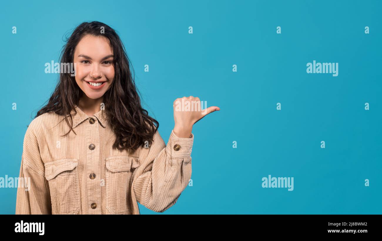 Sorridente giovane donna bruna europea in casual puntando dito verso spazio vuoto isolato su sfondo blu Foto Stock