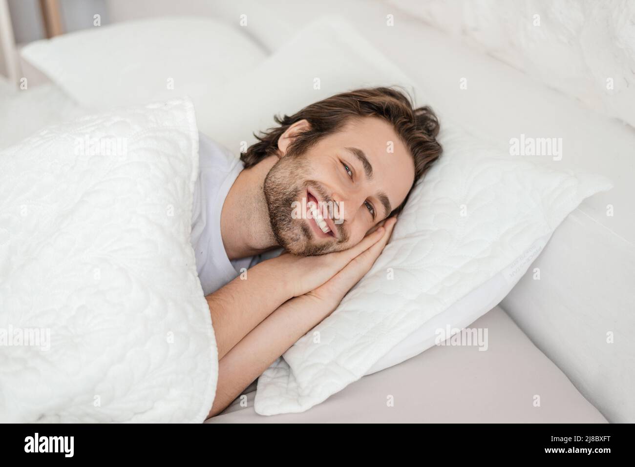Sorridente maschio caucasico millenario con sveglia stoppia, si trova sul letto bianco, gode di tempo libero e fine settimana Foto Stock