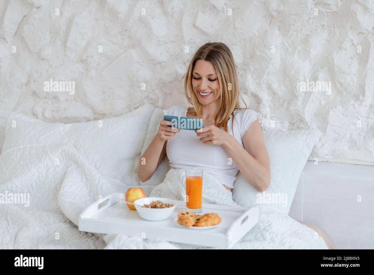 Allegro bella giovane bionda europea donna fa foto di utile prima colazione in letto da solo in camera bianca Foto Stock