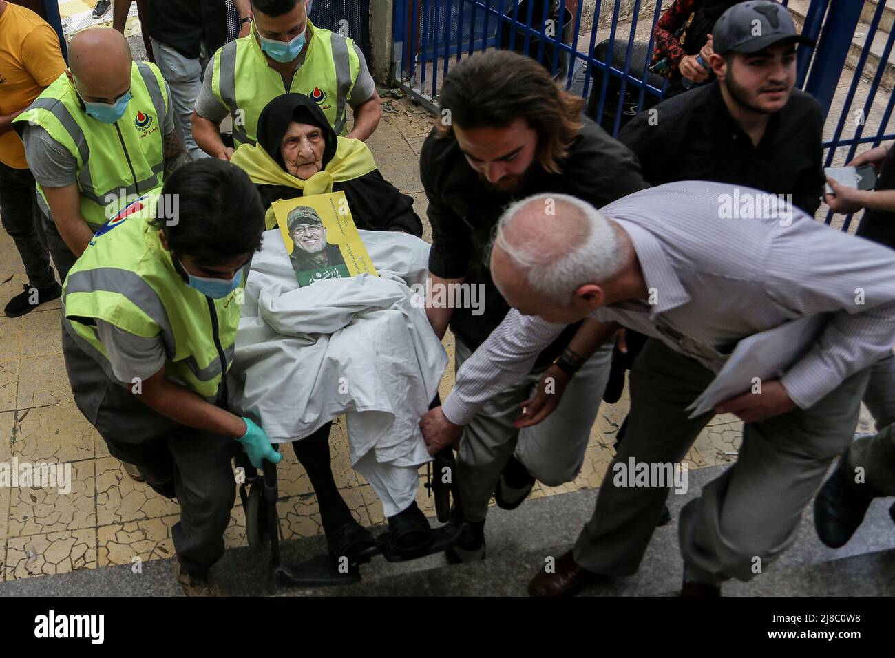 Beirut, Libano. 15 maggio 2022, Libano, Beirut: I membri della difesa civile libanese trasportano Fatima Jezzini, 94, portando la bandiera gialla pro-iraniana di Hezbollah intorno al collo e la foto del capo morto di Hezbollah Mustapha Badreddine, uno dei sospetti di aver complottato l'assassinio del compianto primo ministro libanese Rafic Hariri, Come arriva a lanciare il suo voto all'interno di un seggio a Beirut principalmente musulmano Shiite sud sobborgo durante le elezioni parlamentari libanesi del 2022. Foto: Marwan Naamani/dpa Credit: dpa picture Alliance/Alamy Live News Foto Stock