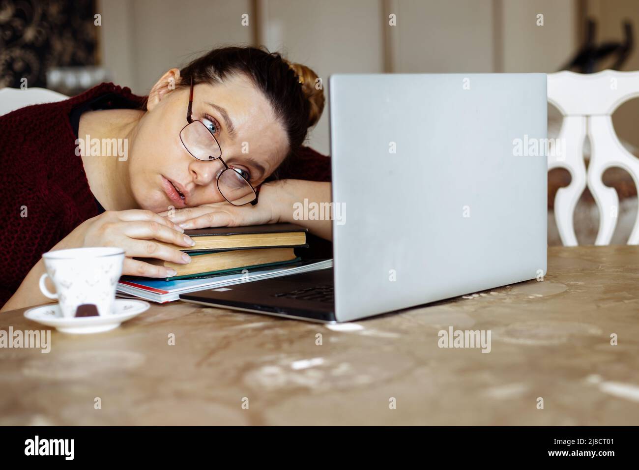 Giovane donna stanca in occhiali sdraiati sui libri e guardando lo schermo del computer portatile alla scrivania della camera di casa. Stanchezza da studio monotono e computer Foto Stock