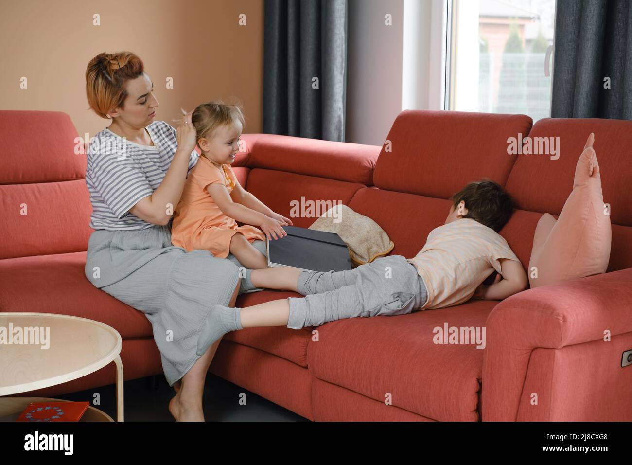 Madre che si prende cura di due bambini a casa sul divano rosso. Fare i capelli per la figlia e parlare con il figlio al mattino. Momento familiare informale. Interazione e. Foto Stock