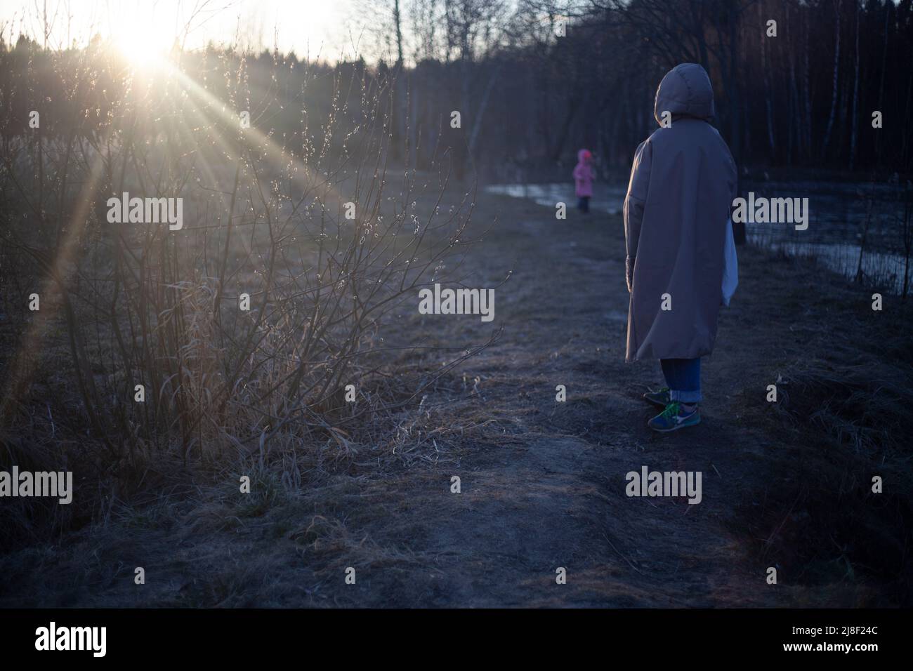 Una ragazza cammina al tramonto con un bambino. Passeggiata sul lago nella foresta. Giovane madre in una giacca calda. Riprese dal retro. Foto Stock
