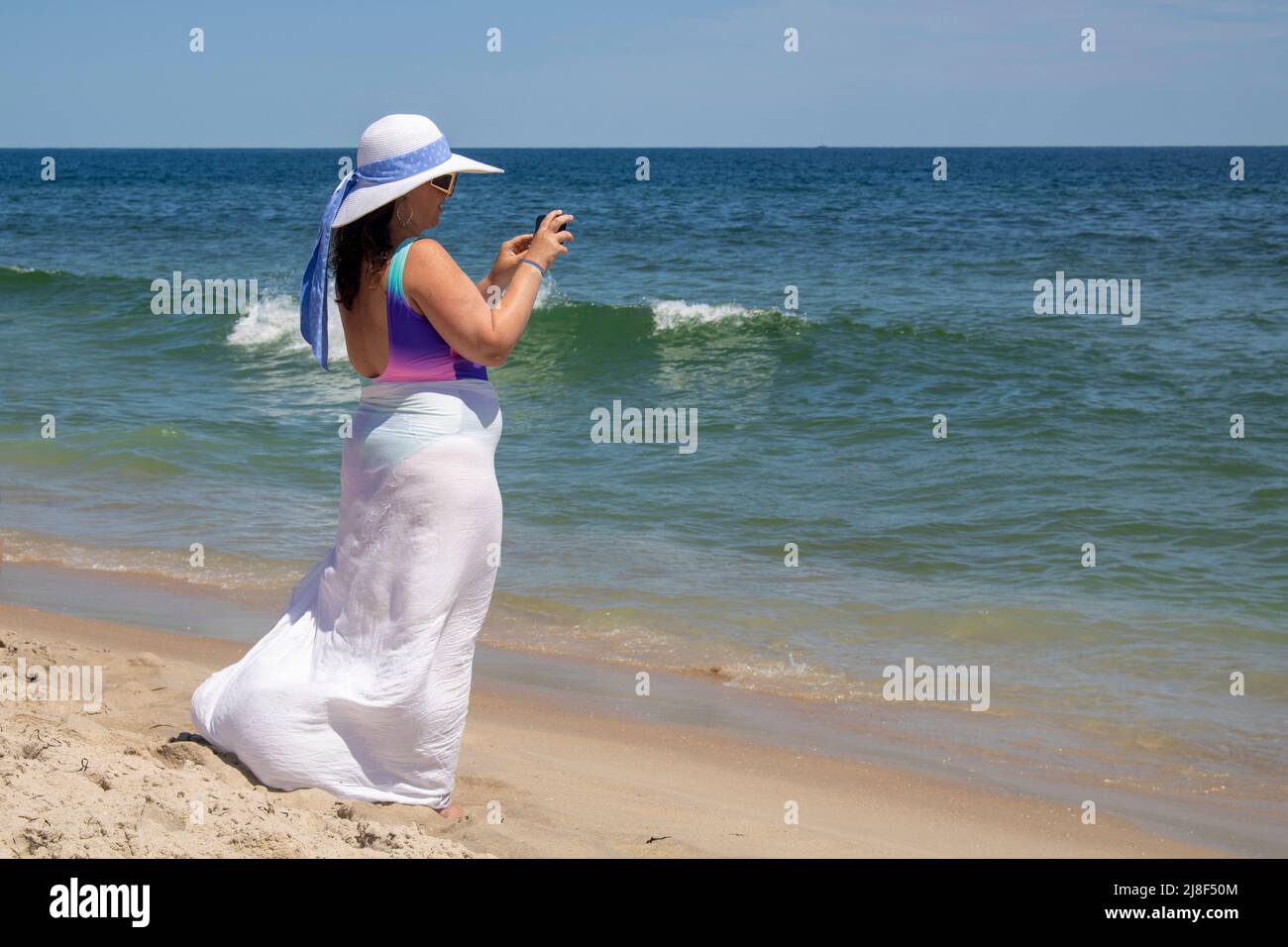 donna chunky con cappello bianco e gonna sopra i bagnanti scattando foto con il telefono delle cellule della spiaggia dell'oceano Foto Stock