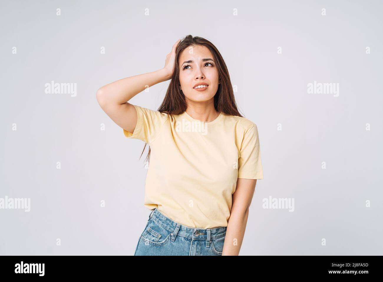 Giovane donna asiatica infelice con capelli lunghi in camicia gialla e jeans che tengono le mani sulla testa su sfondo grigio Foto Stock