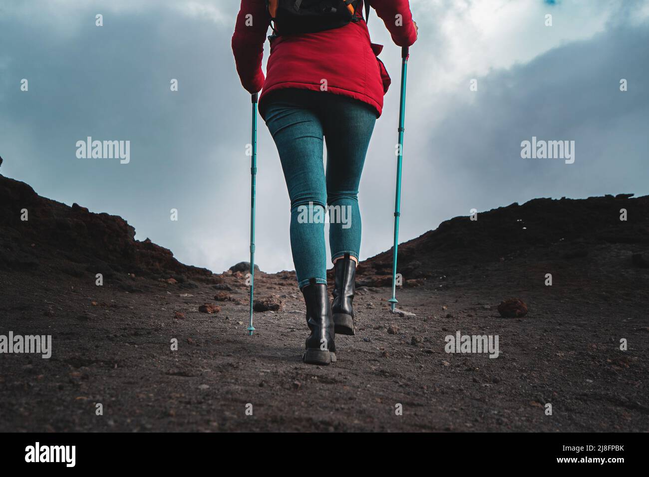 Vista posteriore di una donna escursionista che cammina sulla pietra lavica del Vulcano Etna in Sicilia con bastoni da trekking, gilet rosso e zaino - stile di vita wanderlust c Foto Stock