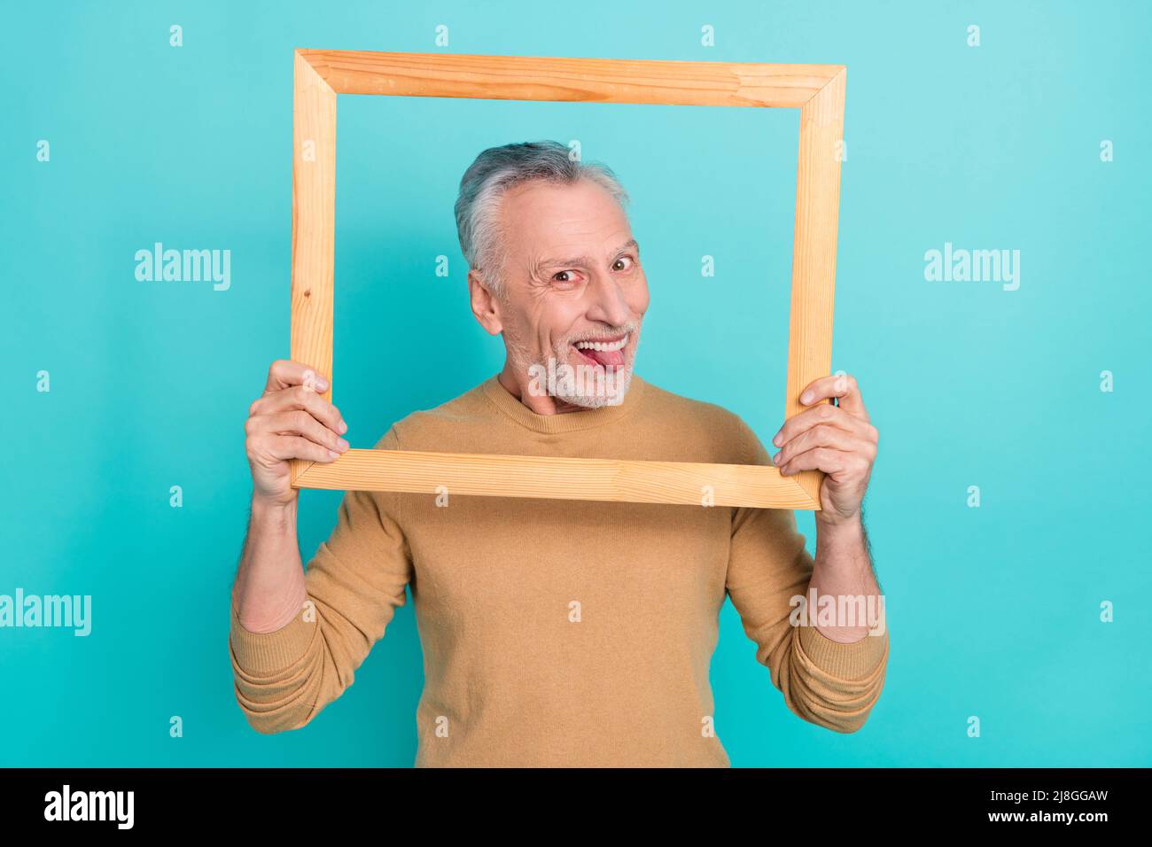 Foto di attraente buon umore maschio foling intorno bastone out lingua tenendo cornice di legno isolato su sfondo di colore teale Foto Stock