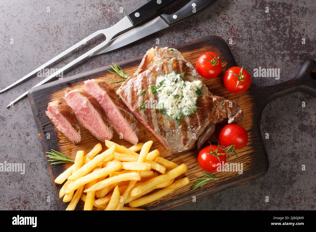 Bistecca di Ribeye alla griglia con fritte francesi, burro verde e pomodori a spicchi sul bordo di legno sul tavolo. Vista dall'alto orizzontale Foto Stock