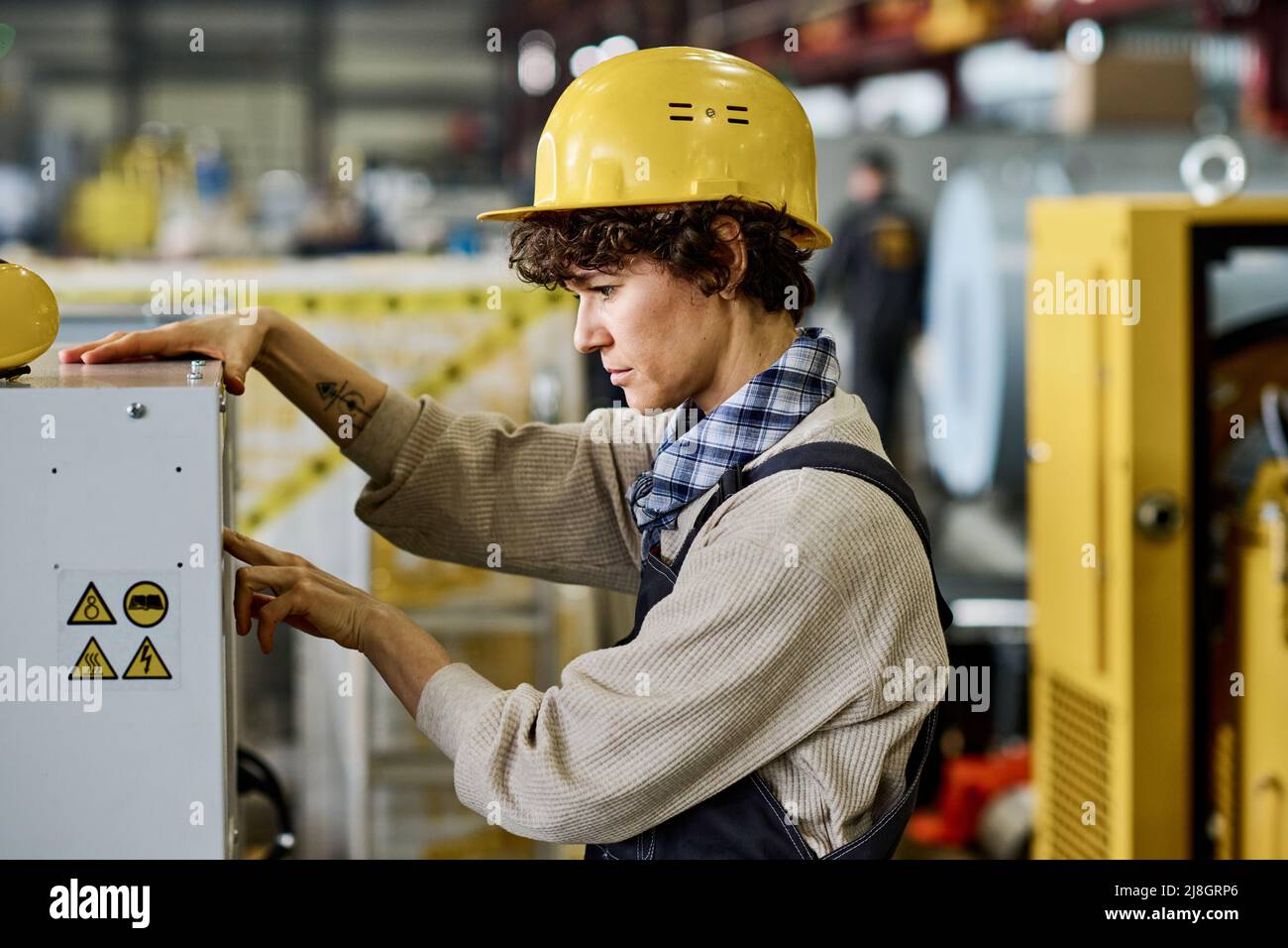 Vista laterale di un ingegnere serio in hardHat premendo il pulsante di avviamento sul pannello della macchina o di altre apparecchiature industriali per controllare come funziona Foto Stock