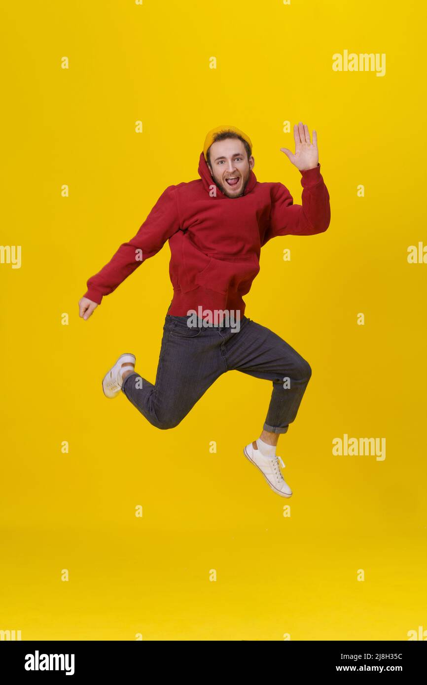 ritratto divertente di un giovane uomo in jumping di gioia alto indossando casual felpa con cappuccio rosso e jeans isolato su sfondo giallo. Hipster humorous ragazzo in salto. Foto Stock