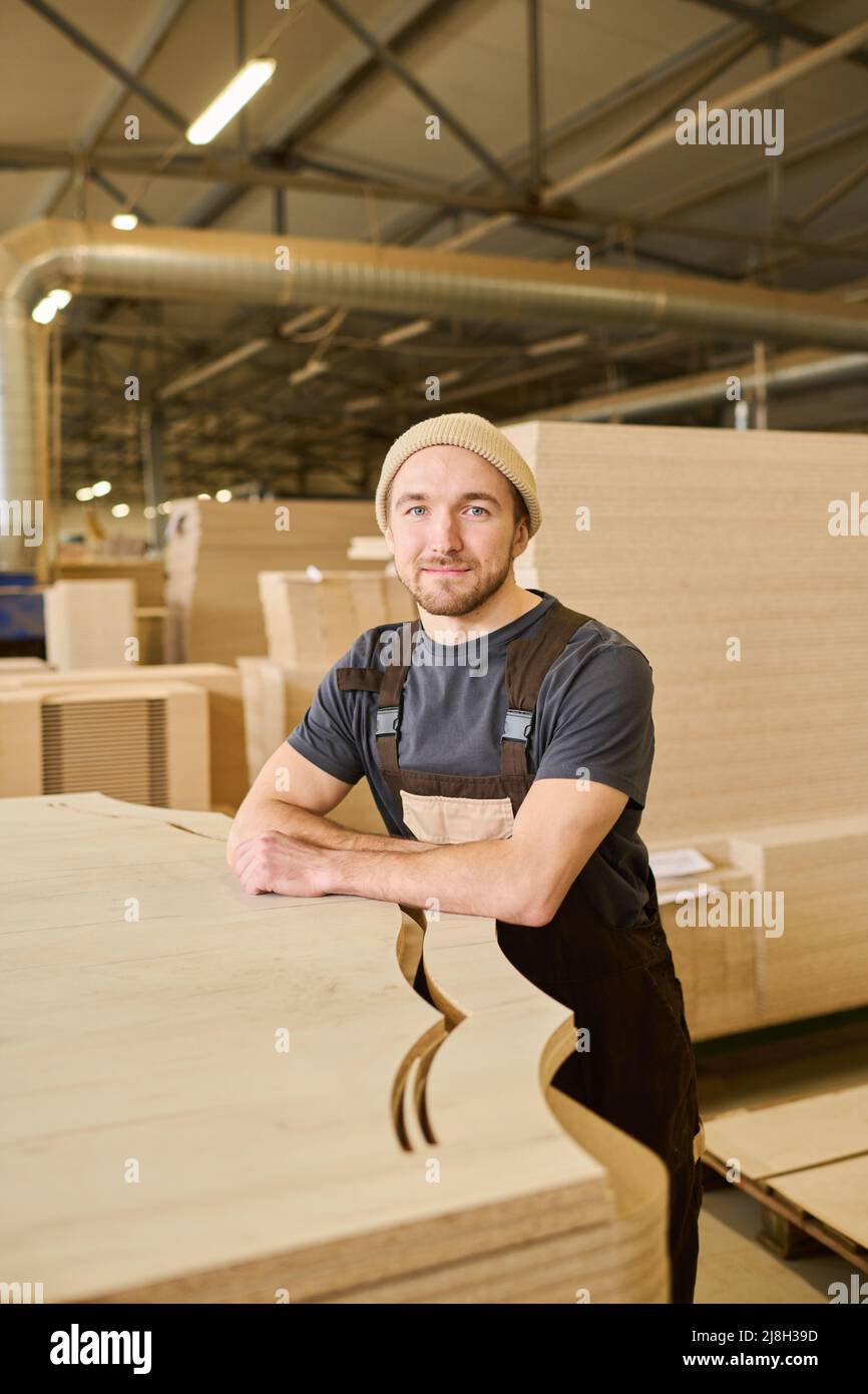 Ritratto di giovane falegname in tute guardando la macchina fotografica in piedi con prodotti finiti in legno presso la fabbrica di mobili Foto Stock