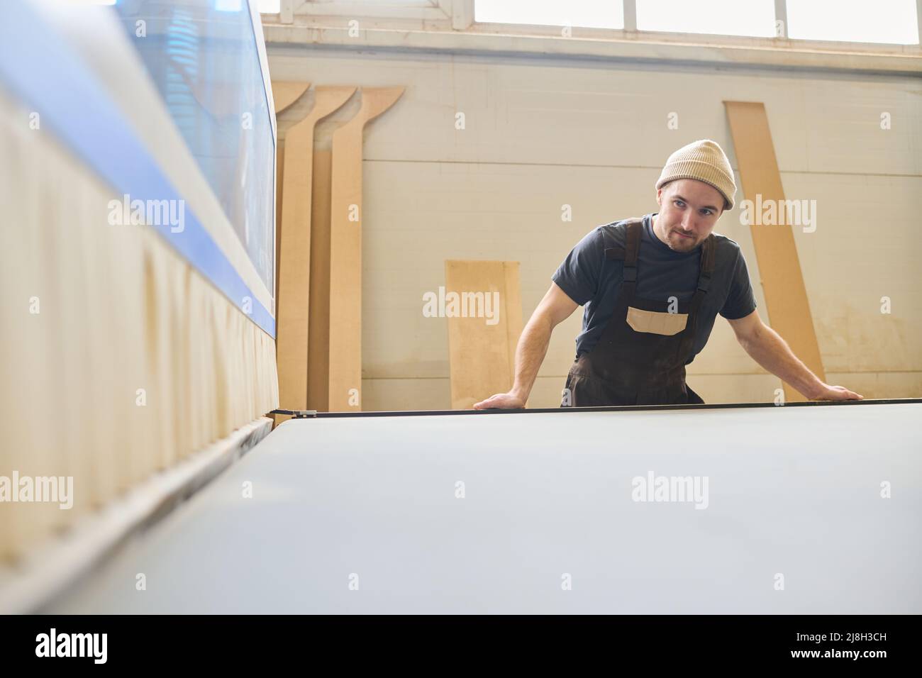 Giovane operaio manuale in lavorazione uniforme a macchina presso la fabbrica di mobili Foto Stock