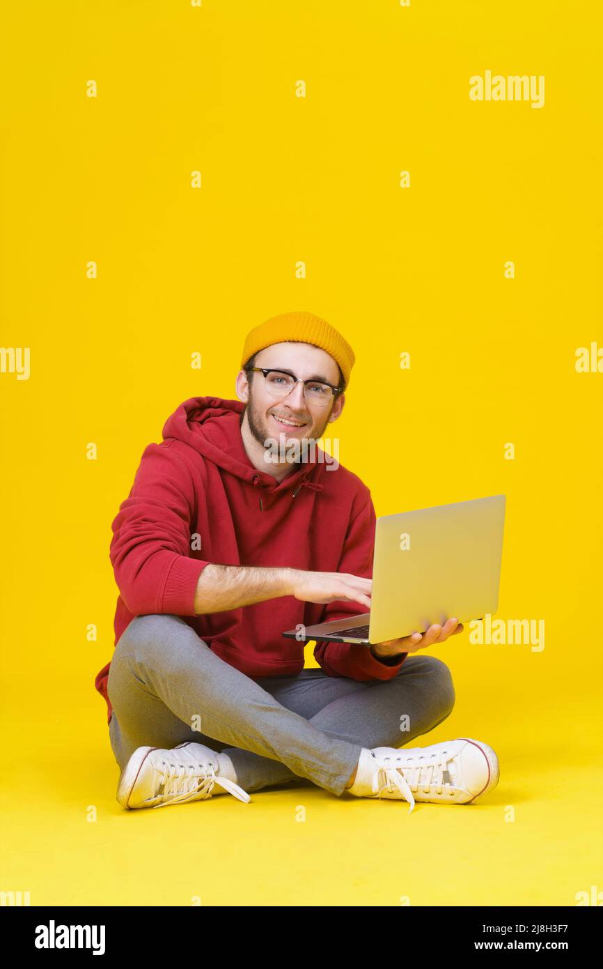 Happy giovane uomo hipster in rosso felpa con cappuccio seduta sul pavimento tenendo un computer portatile a studiare o a lavorare. Uomo caucasico freelance intelligente con notebook isolato su sfondo giallo. Foto Stock