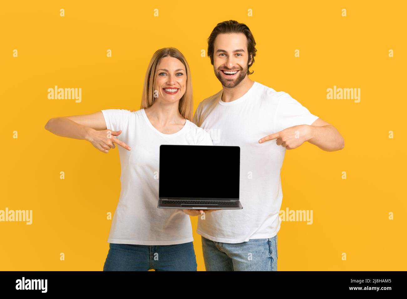 Sorridente coppia europea millenaria in t-shirt bianche puntano le dita al laptop con schermo vuoto Foto Stock