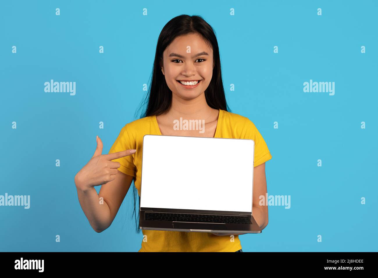 Felice giovane donna giapponese blogger in giallo t-shirt puntino dito al laptop con schermo vuoto, consiglia sito web Foto Stock