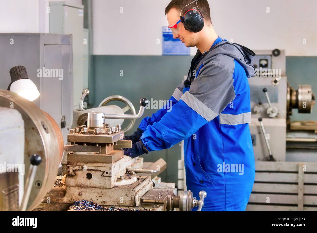 Il giovane turner in occhiali e cuffie di sicurezza lavora al tornio in officina. Flusso di lavoro della scena autentico. L'operatore caucasico elabora le parti metalliche. Foto Stock