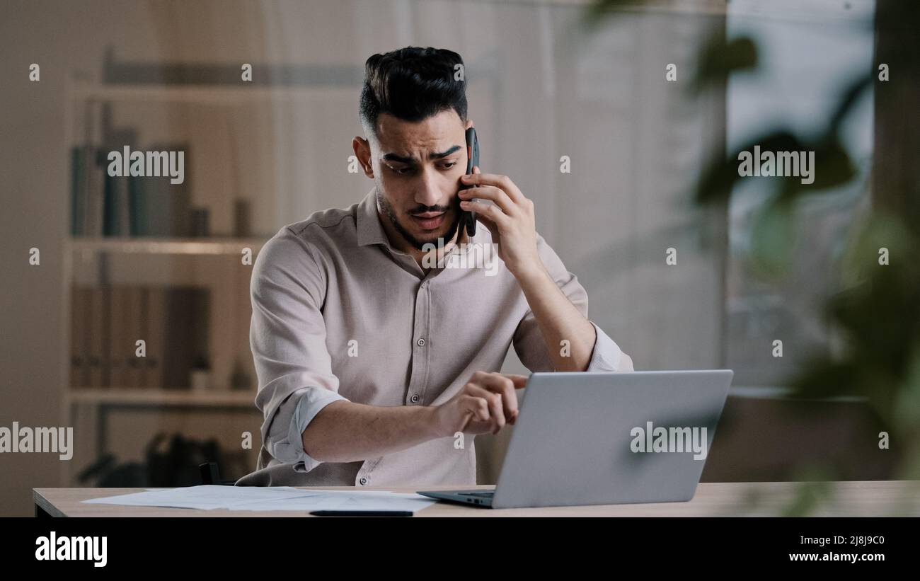 Occupato uomo d'affari arabo millenario dipendente web designer utilizzando laptop parlare sul cellulare coinvolti in multitasking utilizzando gadget discutere online Foto Stock