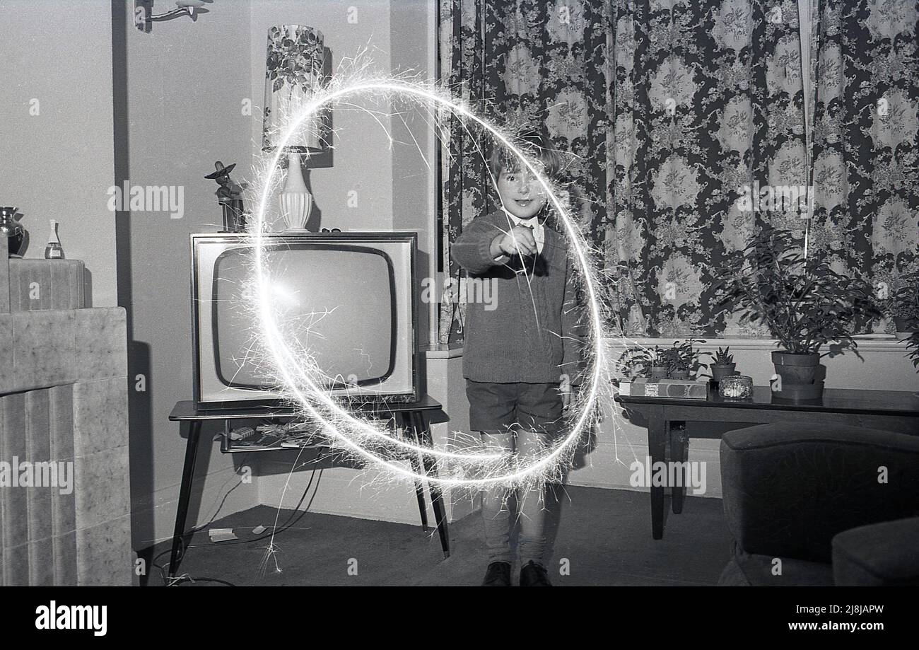 1970s, storico, un giovane schoooboy che gioca al chiuso in una sala con un scintillio di fuochi d'artificio, creando un cerchio di luce che brucia, Inghilterra, Regno Unito. Fuochi d'artificio a mano, gli sparklers sono fatti da un filo metallico con un'estremità coperta in un combustibile di miscela di metallo, che quando acceso produce scintille brucianti lente. Tradizionalmente giocate con i bambini, sotto la supervisione di un adulto, prima di una festa o esposizione più grande dei fuochi d'artificio, gli scintille risalgono all'antica Grecia, dove erano originariamente creati come arma. Foto Stock