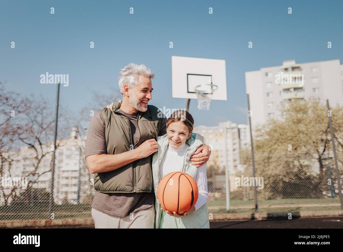 Felice padre e figlia teen abbracciando e guardando la macchina fotografica fuori al campo di pallacanestro. Foto Stock