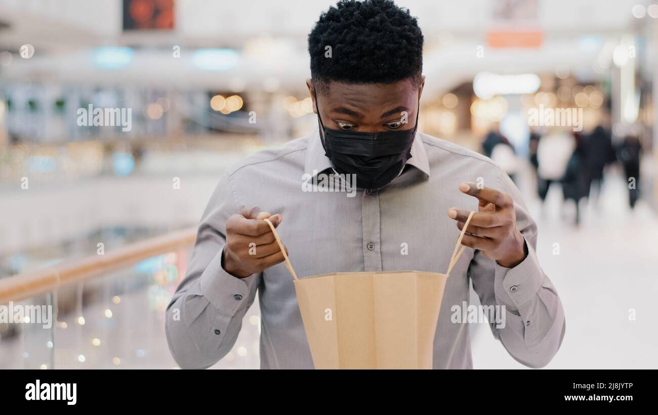 Primo piano giovane africano americano uomo in maschera medica protettiva in piedi in centro commerciale maschio cliente acquirente compleanno uomo guardando nel pacchetto con nuovo acquisto Foto Stock