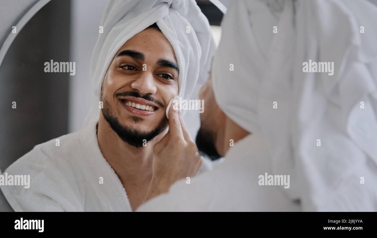 Divertente uomo millenario bello ported indiano ragazzo arabo con asciugamano bianco sulla testa guarda in riflessione in bagno specchio bello-guardando maschio a casa Foto Stock
