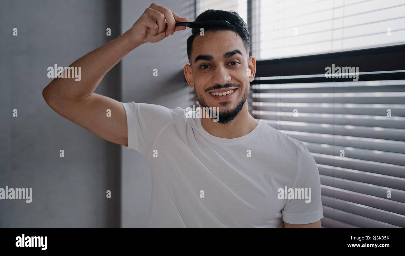 Arabian spaniard arab uomo che guarda la macchina fotografica pettinare i capelli con una spazzola che si prepara ad oggi lavoro giorno a casa ragazzo in bianco T-shirt mattina bellezza Foto Stock
