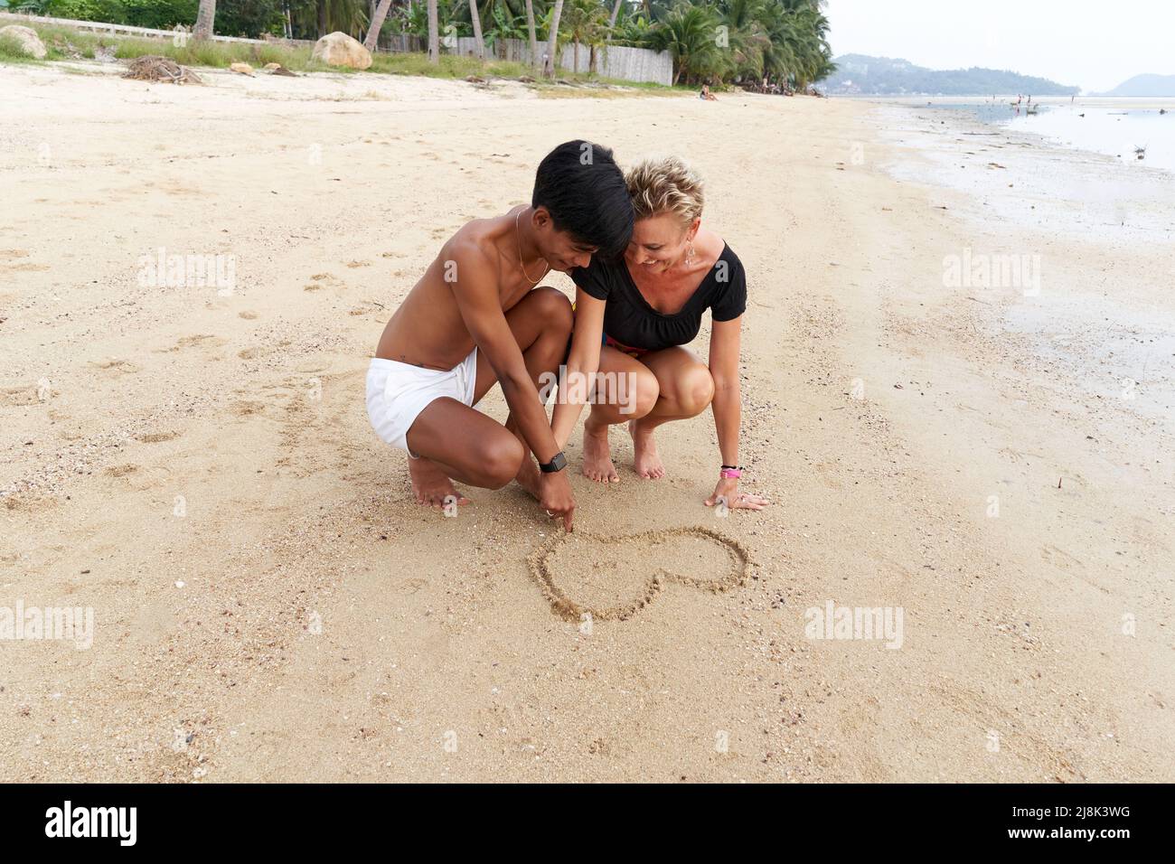 Coppia multiculturale su una spiaggia che disegnano un cuore forma insieme sulla sabbia Foto Stock