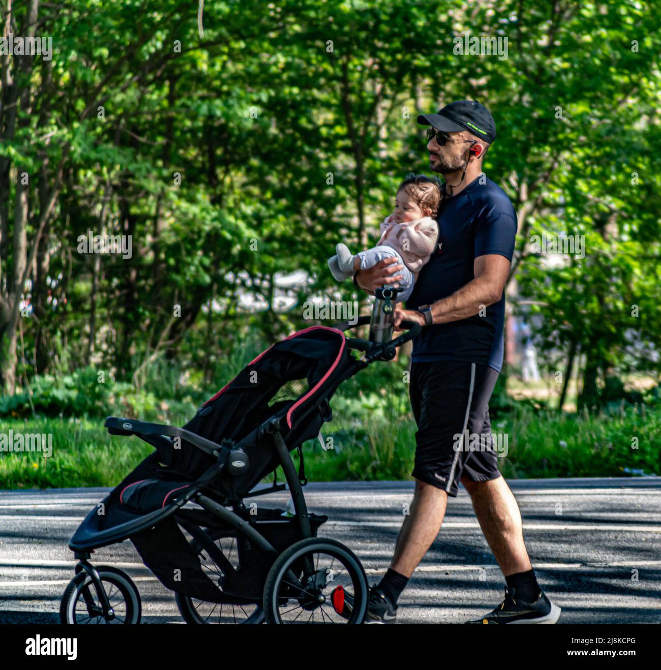 Un uomo sta tenendo un bambino in una mano e passeggino in un altro esercizio nel parco nel caldo giorno della primavera del 2022 Foto Stock