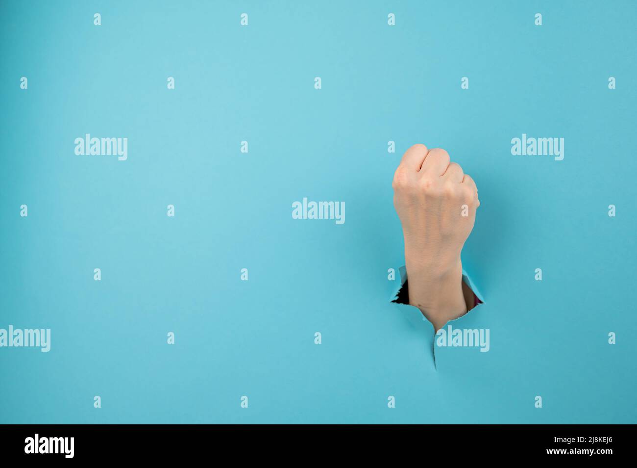 La mano di una donna che sporge da un buco da uno sfondo blu mostra un pugno. Foto Stock