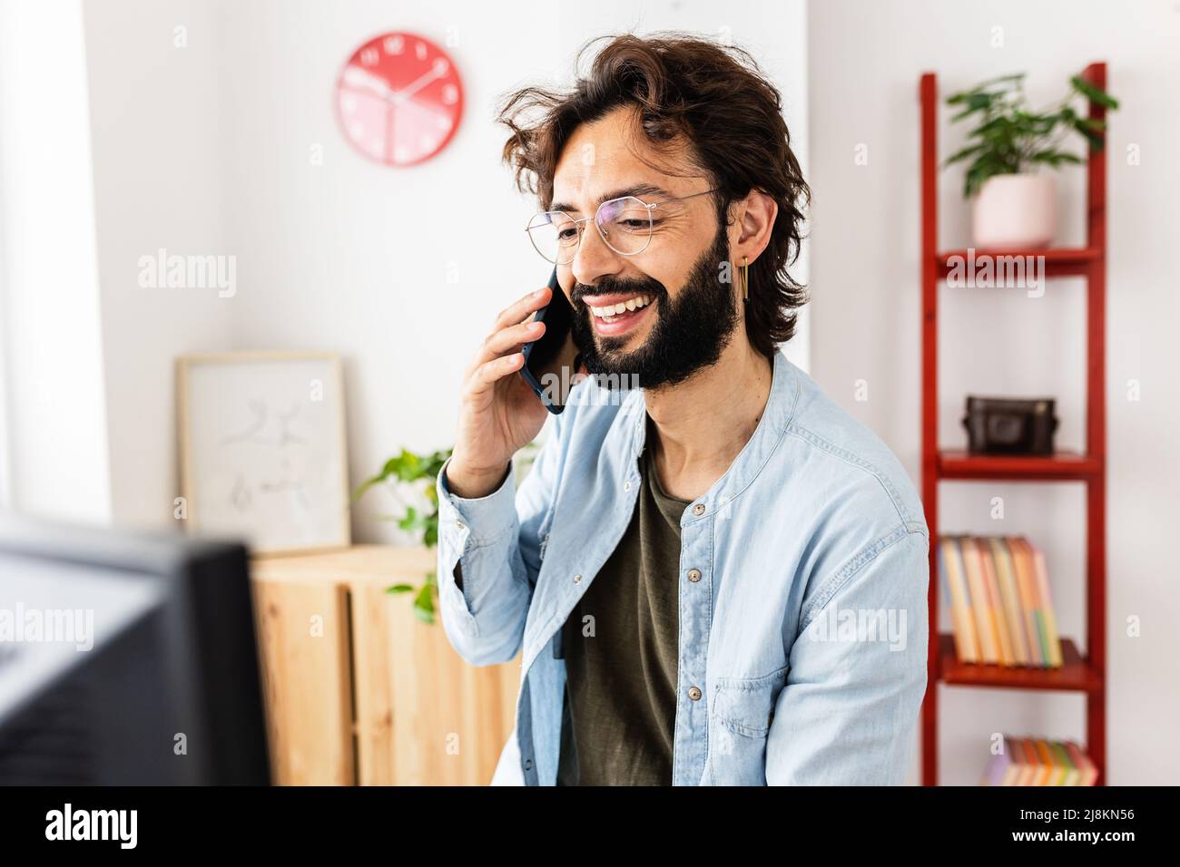 Uomo sorridente che parla al telefono mentre lavora a casa. Foto Stock
