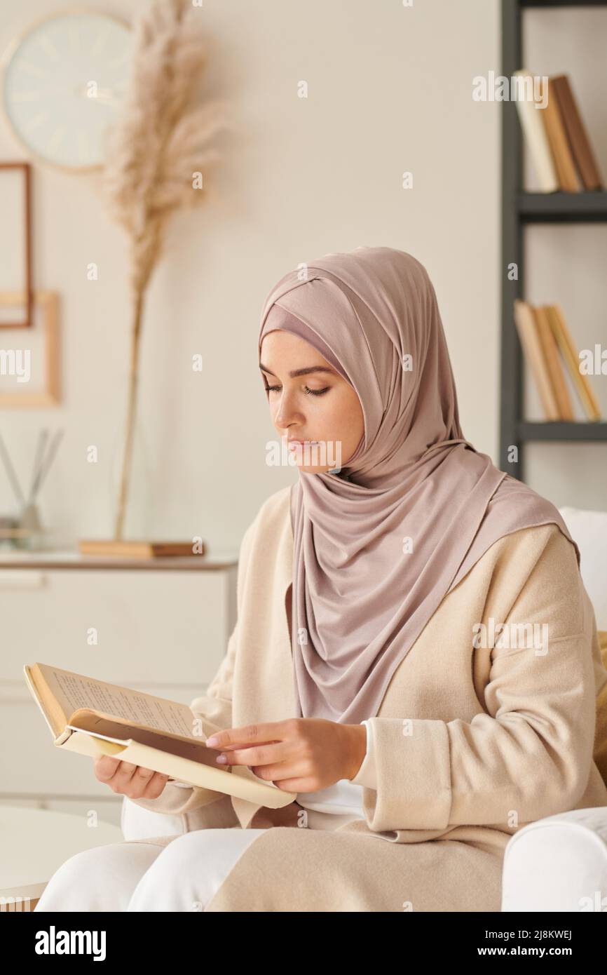 Bella giovane donna che indossa elegante hijab rosa pallido seduta in poltrona da solo in soggiorno lettura romanzo Foto Stock