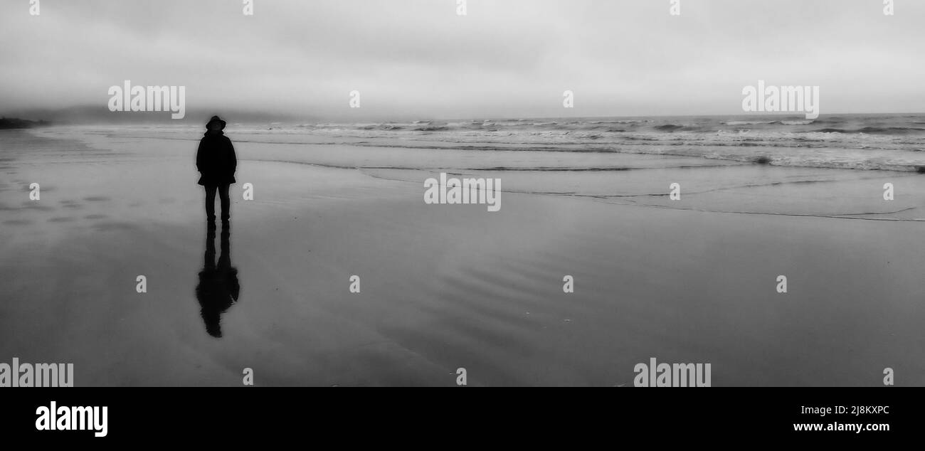 In un'immagine sfocata, una figura senza volto rivestita di nero si erge su un'ampia spiaggia sabbiosa della Nuova Zelanda , in stile Michael Orton Foto Stock