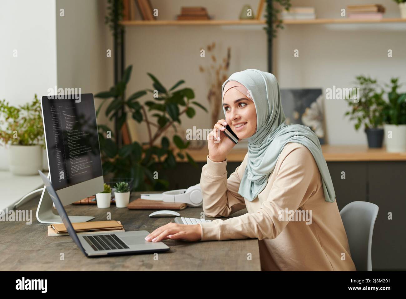 Ragazza allegra che indossa un hijab azzurro seduto al tavolo che parla al telefono mentre lavora al computer Foto Stock