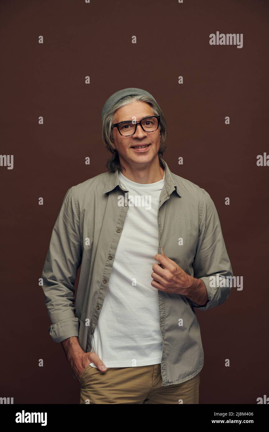 Ritratto di uomo maturo serio e di buona qualità con capelli grigi che indossano cappello da hipter su sfondo marrone Foto Stock