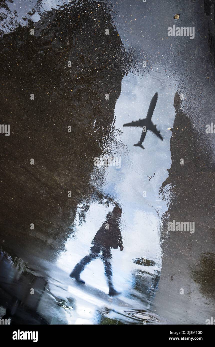 La silhouette sfocata del riflesso di una persona che cammina da sola sul marciapiede bagnato della città nelle giornate piovose. L'aereo vola attraverso il cielo. Fotografia astratta. Foto Stock