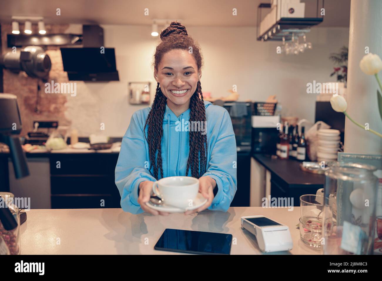 Bella giovane barista femminile di razza mista con trecce che consegna tazza di caffè alla macchina fotografica. Foto Stock