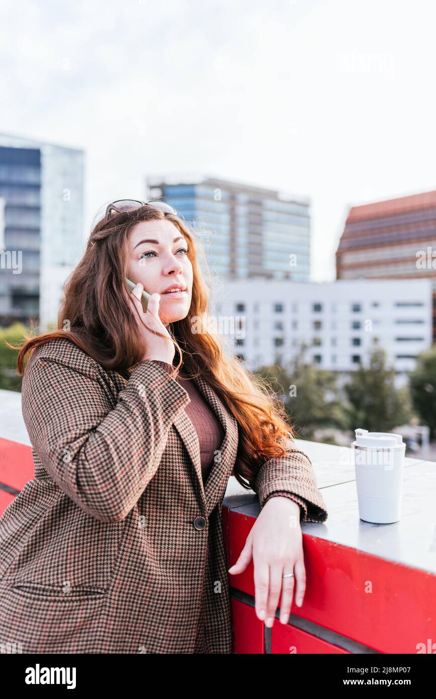 Donna in cappotto a scacchi con capelli lunghi appoggiati sul bordo del ponte vicino al caffè per andare. Sta guardando lontano mentre parla al telefono cellulare sulla strada della città moderna. Foto Stock