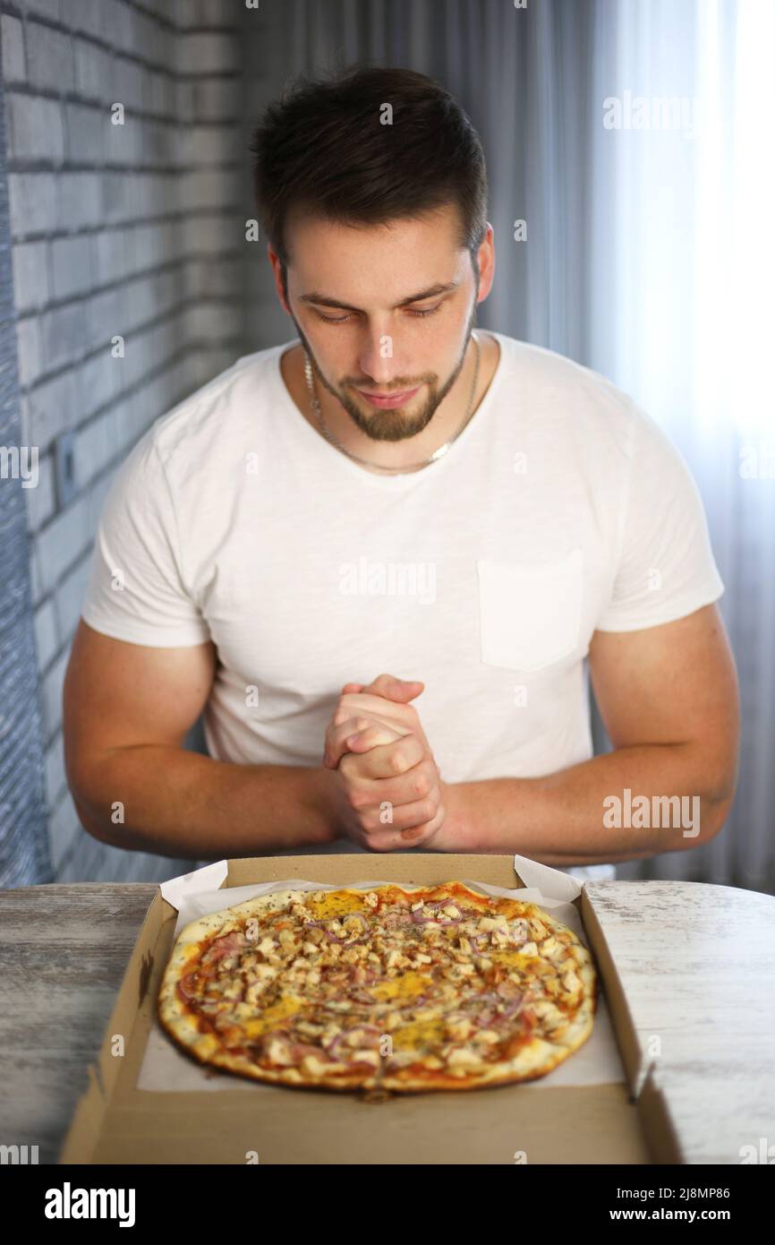 Un giovane atletico in una t-shirt bianca si siede ad un tavolo e guarda un pacchetto aperto di pizza Foto Stock