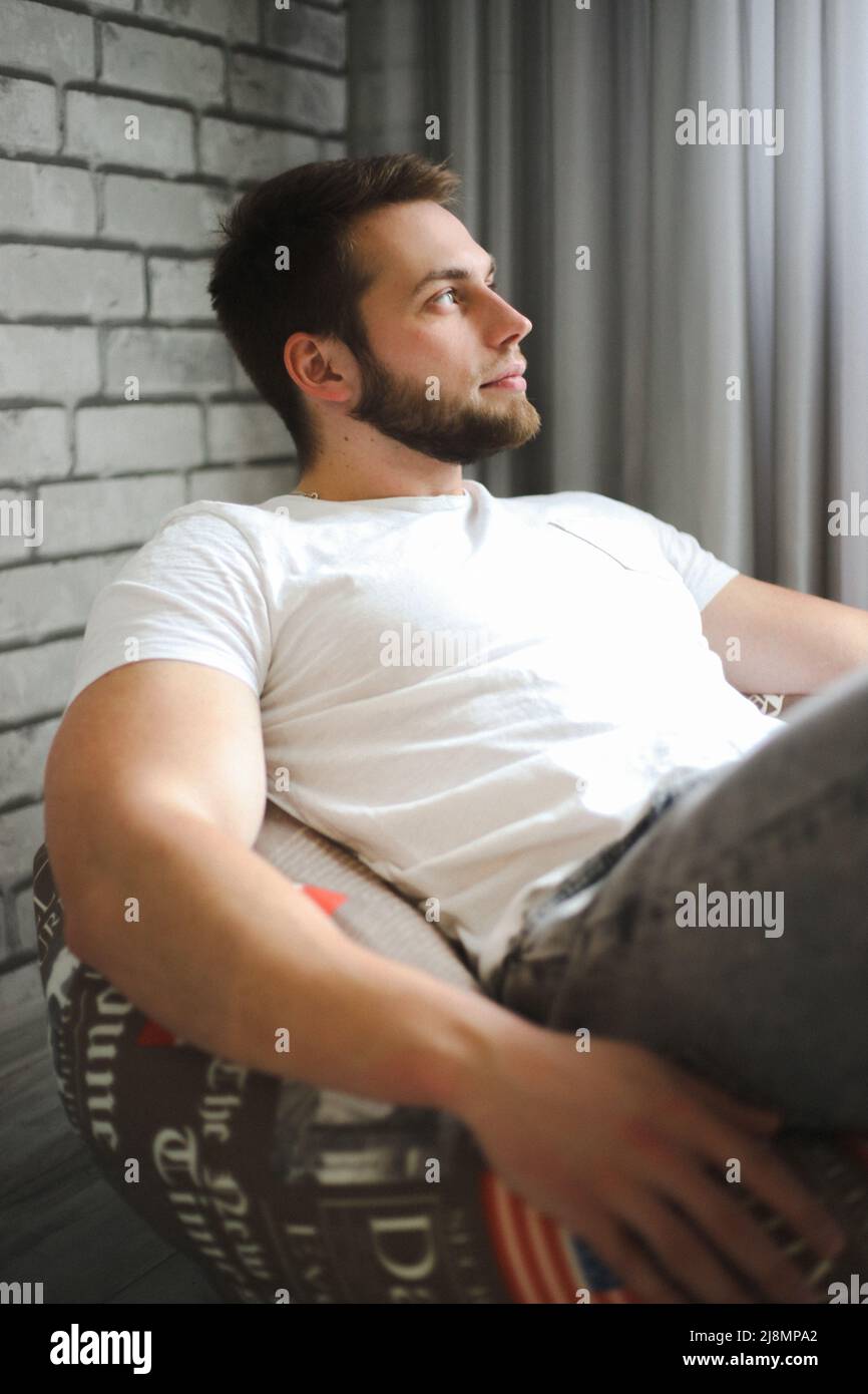 Un giovane uomo di atletica in una T-shirt bianca si siede su un cuscino di poltrona e guarda con attenzione la luce Foto Stock