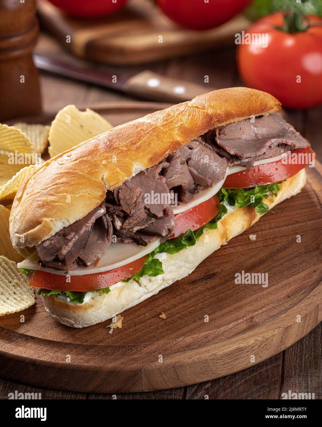 Sandwich sottomarino con roast beef, formaggio, pomodoro e lattuga su un rotolo di hoagie con patatine fritte su un tagliere Foto Stock