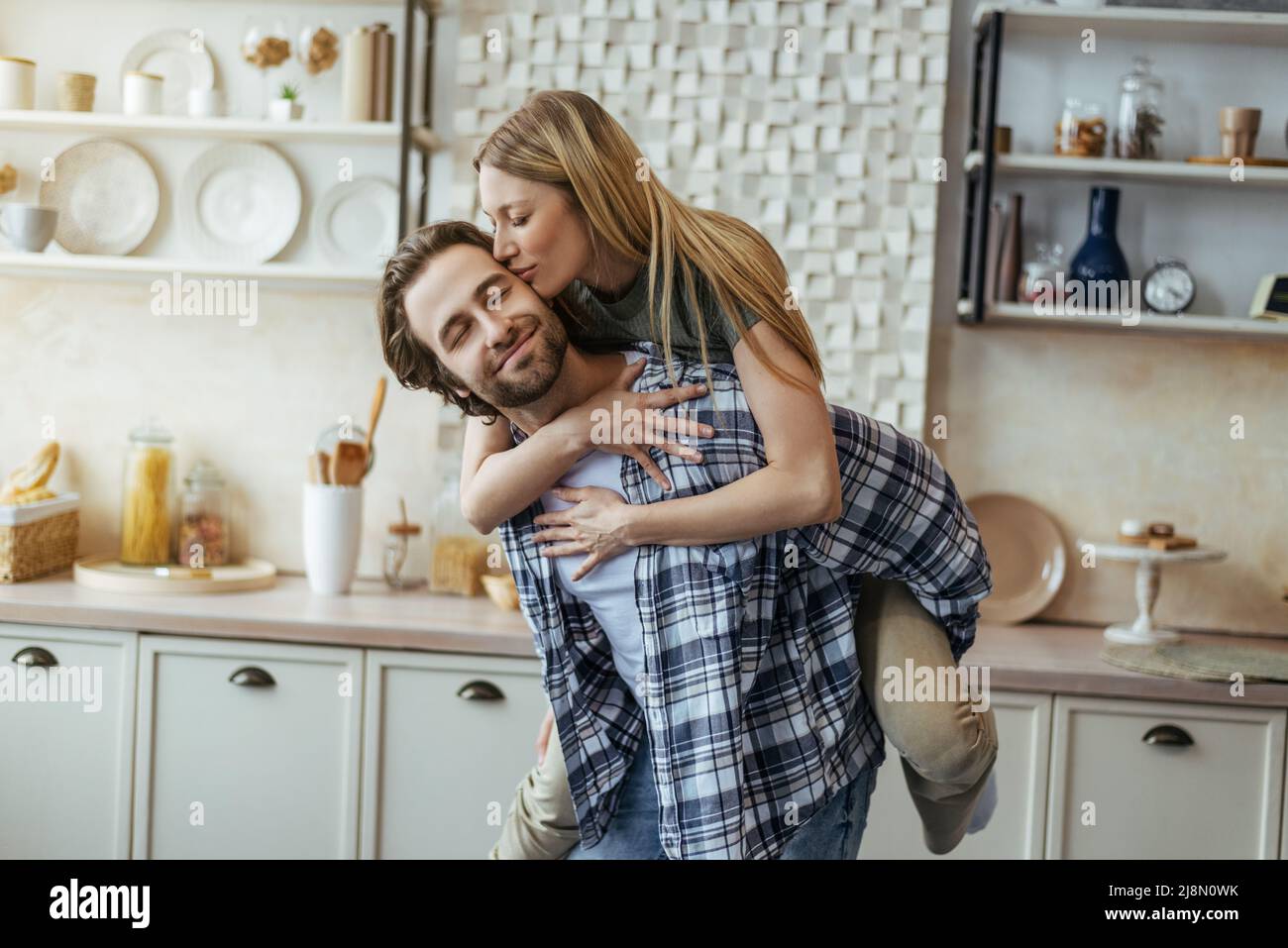 Soddisfatto ragazzo millenario europeo con stoppia tiene la donna bionda sulla sua schiena, signora bacia guancia, godere del tempo libero Foto Stock