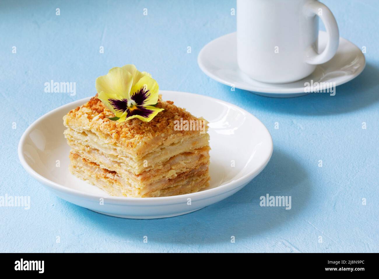 Una fetta di torta Napoleone, decorata con un fiore di viola, servita con una tazza di caffè o tè. Foto Stock
